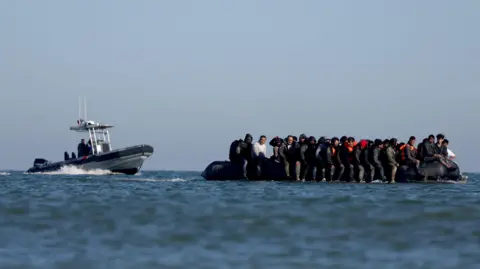 Reuters A group of migrants, some of them wearing safety vests, are sitting on an inflatable dinghy at sea. A French police boat is approaching them from behind. The sky is blue and the sea is relatively calm.