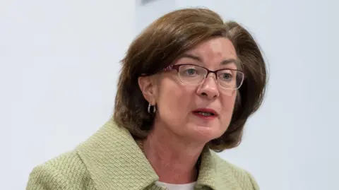 Senedd Cymru Eluned Morgan stood in the temporary Senedd debating chamber, wearing a green jacket and glasses. She has dark brown shoulder-length hair