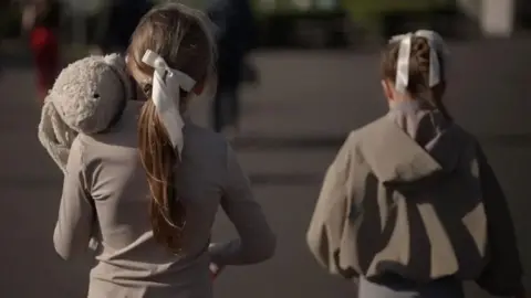 Two girls wearing grey tops and white ribbons in their hair face away from the camera. One is carrying an off-white teddy bear