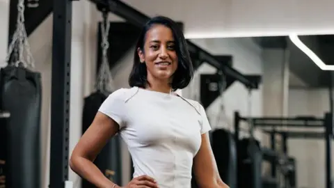 Samina Zaman Samina Zaman has shoulder length black hair, a white top and is smiling. There is gym equipment behind her.