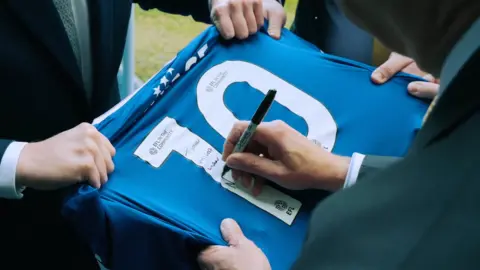 Reform UK A still from a video showing a hand signing the back of a blue football shirt that has the number 10 on it.