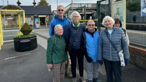 Zes leden van de Friday Strollers glimlachen naar de camera tegen de achtergrond van het treinstation van Maghull