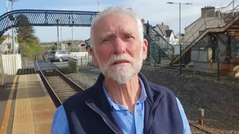 A man with balding white hair and a white beard. He is wearing a blue shirt and a sleeveless navy fleece. There is a train track behind him and a black bridge above it. 