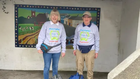 Kerstin Banham Mr and Mrs Banham wearing their grey Pilgrimage sweatshirts and belt packs. Mr Banham is wearing a brown cap. They are standing in front of a mural showing a village scene with a church, houses, cows and a river.