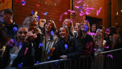 A queue outside a nightclub, with several people waiting to get in and dressed up for a night out. Confetti is being flung in the air as they wait.