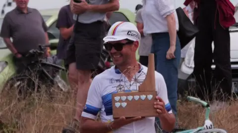 Alexandre Voisine in cycling clothing and a cap, holding a wooden trophy depicting a penny farthing and smiling.