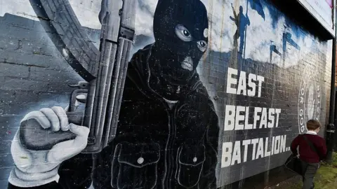 Mural of a man wearing a balaclava and holding a gun, writing beside it says East Belfast Battalion and a pupil is visible walking beside it