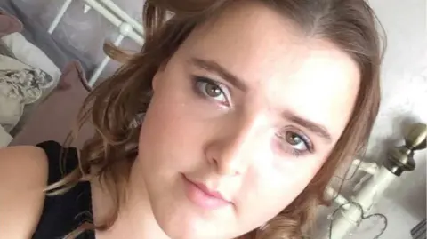 A young woman sitting on a bed with her hair curled and make-up on, looking at the camera in selfie-style image.