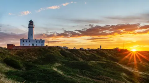 Chris McLoughlin / Getty Images An orange sunset over grass verges. A white lighthouse is on the left.