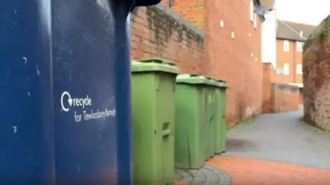 A photo of a row of blue and green bins located on a street in Tewkesbury next to a red bricked property.
