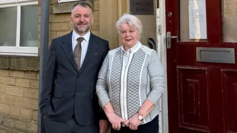 Angela Viney Angela Viney on the right and Darren Becks, both dressed in smart clothes as they stand in a paved street in front of a dark red door.
