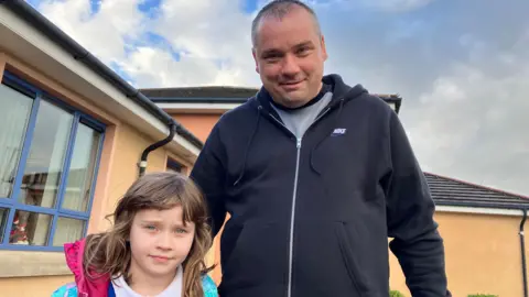 a man and a little girl are pictured outside a school building at Bready primary School. The little girl is on the left, and has long brown hair. The man is wearing a blue hoodie xipped up