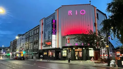 Rio Cinema Rio Cinema sign illuminated in purple with red hue on white building