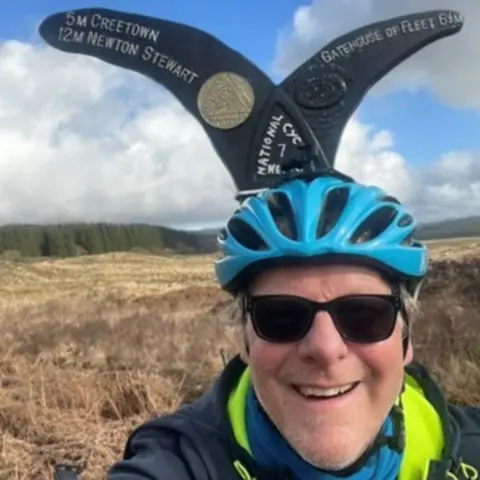 Mike Blakeman A man in a blue cycling helmet wearing sunglasses and a jacket stands in front of a a directions sign with grassland behind him and trees in the distance