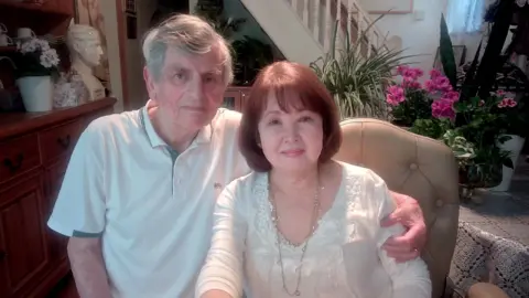 A man and a woman sat side by side on chairs. She has auburn hair to her shoulders and wears a white top with a silver necklace. He has a white polo shirt on and short, grey hair.