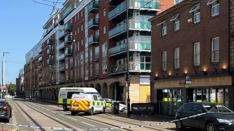 Oli Constable/BBC Blue and white police tape stretches across a city centre street. behind the tape, two police vans and a police car are parked , blocking the junction to a side street. The bar is a three-storey red brick building with floor to ceiling windows at ground level and two rows of four standard windows on the floors above. The next building along the street is a modern seven-storey block of apartments with glass-fronted balconies. It is a sunny day with a blue sky.