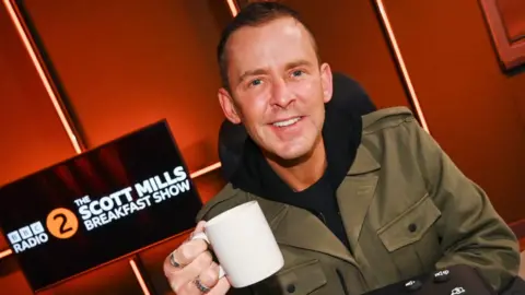 Scott Mills holding up a coffee mug, in the Radio 2 studio