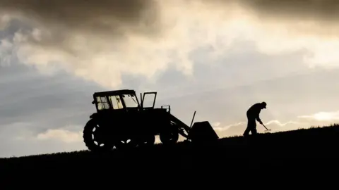 A tractor and farmer raking the ground are silhouetted against a blue sky with yellow clouds.