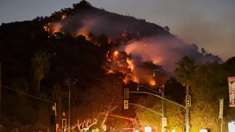 Getty Images A forested hill rising above a street at night. The trees are on fire with smoke billowing into the sky.