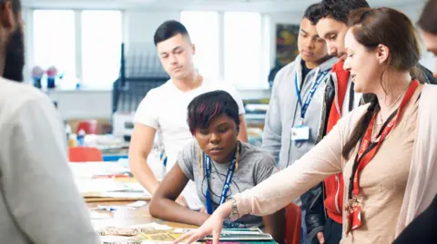 Getty Images A lecturer in a classroom is pointing to a desk with different fabrics on, surrounded by a small group of students in a textiles class. They all have lanyards on their necks - the lecturer's is red and says "staff". One female student is sat at the desk close to where the lecturer is pointing, while two other male students are stood behind her.
