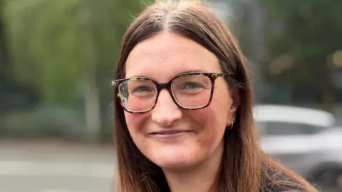 Gemma Dillon/BBC The image shows a lady wearing glasses. She has long brown hair and is smiling.