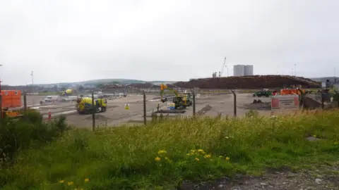 A site at the Port of Workington, which is being used for the development of a facility to support the decommissioning of a nuclear reactor in Japan. The site is fenced off and there are heavy plant machines on the concreted yard. In the distance there are cranes and large tanks.