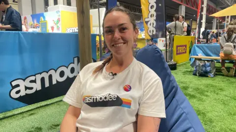 A woman with brown hair, tied back in a plait, and wearing a white shirt with the 'asmodee' logo, smiles as she sits down on a blue seat in the middle of an exhibition hall.