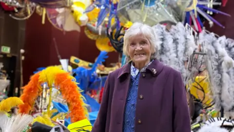 Danny Fullbrook/BBC A woman in a long purple coat is standing in a large room full of carnival costumes, floor to ceiling.