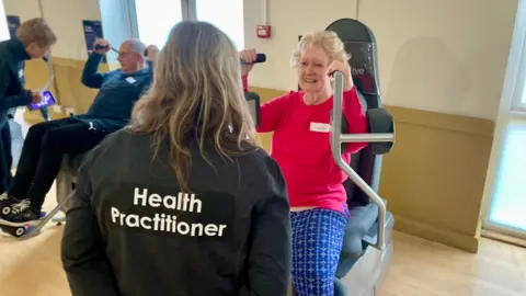 Mark Norman/BBC A woman with back to us with the words 'Health Practitioner' on her sweat shirt and she is standing in front of an older woman who is sitting on a piece of rehabilitation equipment used to exercise her arms. 