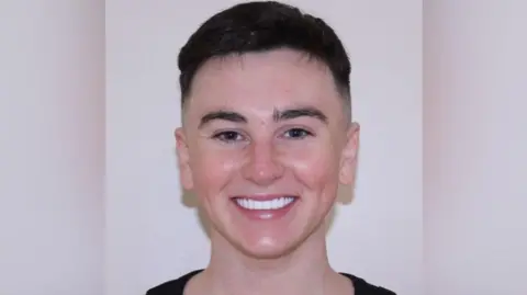 Joshua wearing a black top and smiling at the camera. He has short dark brown hair. He is standing in front of a white wall. 