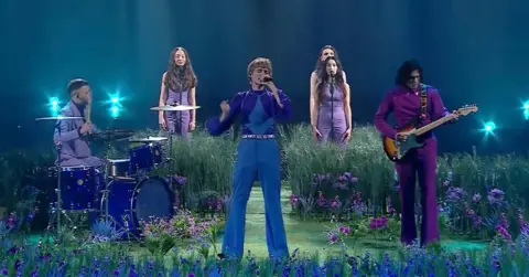 Suspilne Ziferblat perform at the national final of Ukraine's Eurovision selection contest. The band are dressed in shades of blue and purple, and are surrounded by flowers and grass.