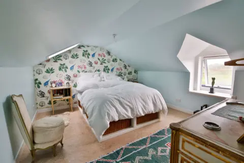 IWC Media A bright attic bedroom features a low, sloping ceiling painted pale blue and a bed dressed in crisp white bedding.
A playful patterned wall with plants and animals, along with small furniture and a window, adds charm and character to the cosy space.