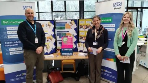 Three NHS staff stand in front of a NHS Devon stand. The workers are wearing lanyard and are standing with their arms by their sides. Two blue and white NHS Devon signs are behind them. A table displaying NHS Devon information has been set up behind them. A sign reads "get involved".