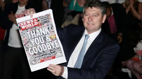 PA Media Colin Myler, the last editor of the News of the World, holds up its last edition as the paper was closed down in 2011. The paper front page says: Thank You & Goodbye. He is wearing a dark suit jacket, and a white shirt with a pale blue tie.