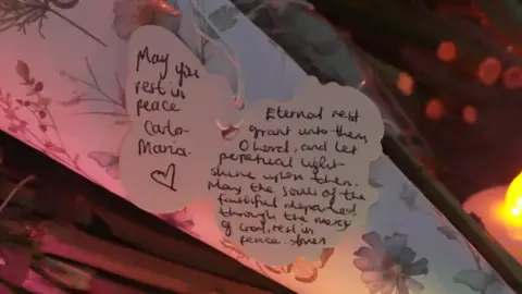 Una foto de cerca de una etiqueta en un ramo de flores, que dice "Que descanses en paz Carla-Maria".