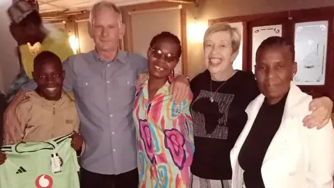 UWE (From left to right) Choir member Muhle, Norman, Muhle's mum Lwazilenkosi Philanathi, Bridget, and Muhle's grandma Zanele during a trip to South Africa. They're all standing next to one another with their arms over each other's shoulders in a line.