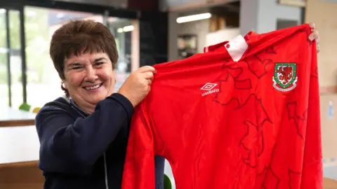 Michelle holds up her old red football jersey with long sleeves, an Umbro logo and a shield with the Welsh dragon on. She smiles at the camera widely. She has brown short hair.