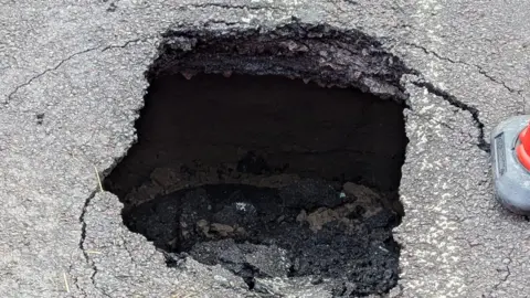 Mike Fielding A close up of the sinkhole shows the collapsed road surface inside the hole surrounded by cracked tarmac and a traffic cone.