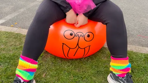 The lower half of a person sitting on an orange space hopper decorated with a silly face. The person is wearing black leggings and a pair of brightly coloured ankle warmers.