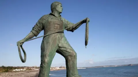 A bronze statue of a fisherman looking out to sea . He holds a rope and wears a beanie type hat. St Michael's Mount is visible across the bay