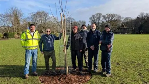 Somerset Council The Mayor of Taunton stands with three other people in a park as she digs into the earth to plant a wild cherry tree