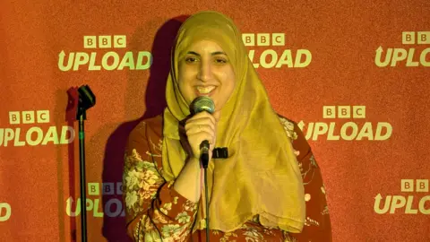 A woman in a gold hijab and red floral shirt holds a microphone to her mouth and smiles. She stands in front of a red backdrop which says "BBC Upload" on it.