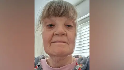 Supplied Mazel Bone has a greying hair tied at the back. She has a fringe and is wearing a pink top and grey sweatshirt. 