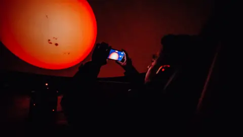 A man, sat looking at his phone, with headphones on, inside a planetarium, with a big orange planet in front of him, that is also on his phone. He is in a chair, half lying back. 