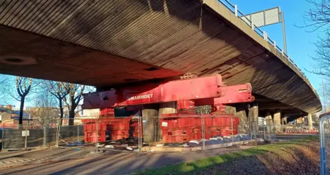 A temporary prop underneath the Gateshead Flyover. It consists of a red metal structure. It is fenced off. The flyover above is made from concrete.