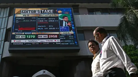 AFP Pedestrians walk past a digital broadcast in front of the Bombay Stock Exchange (BSE) in Mumbai on November 21, 2024. Shares of Indian conglomerate Adani plunged on November 21 after industrialist Gautam Adani was charged by US prosecutors with money laundering. $250 million in bribes for key contracts.