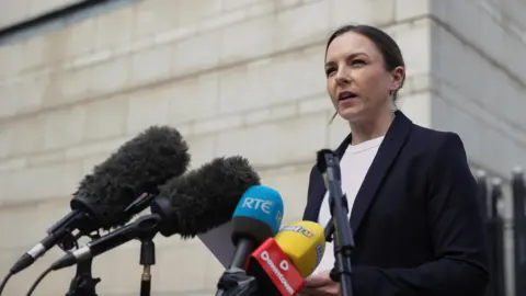 Liam McBurney/PA Wire Det Insp Kelly Foley, a woman with short, dark hair tied back from her face, addresses reporters outside the Laganside court complex in Belfast. She is wearing a dark suit and a white crew-neck top. 