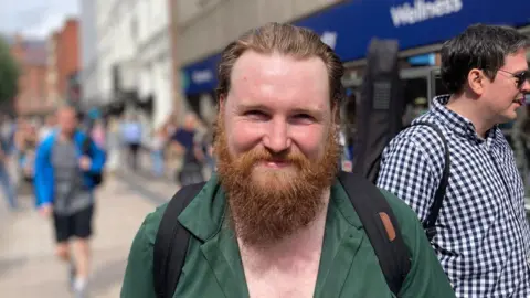 A man smiles at the camera. He has an auburn beard and is wearing a green shirt. He is standing on a busy street. 