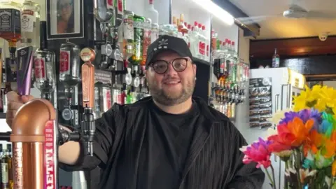 Jenkin Edwards A man with a beard, glasses and a cap stands behind the bar in a pub
