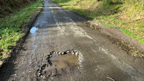 BBC A narrow country road with grass verges on either side and a large pothole filled with water in the foreground